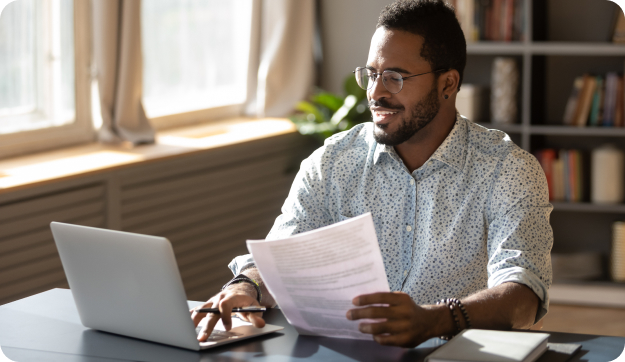 A man browsing a laptop and reading a piece of paper